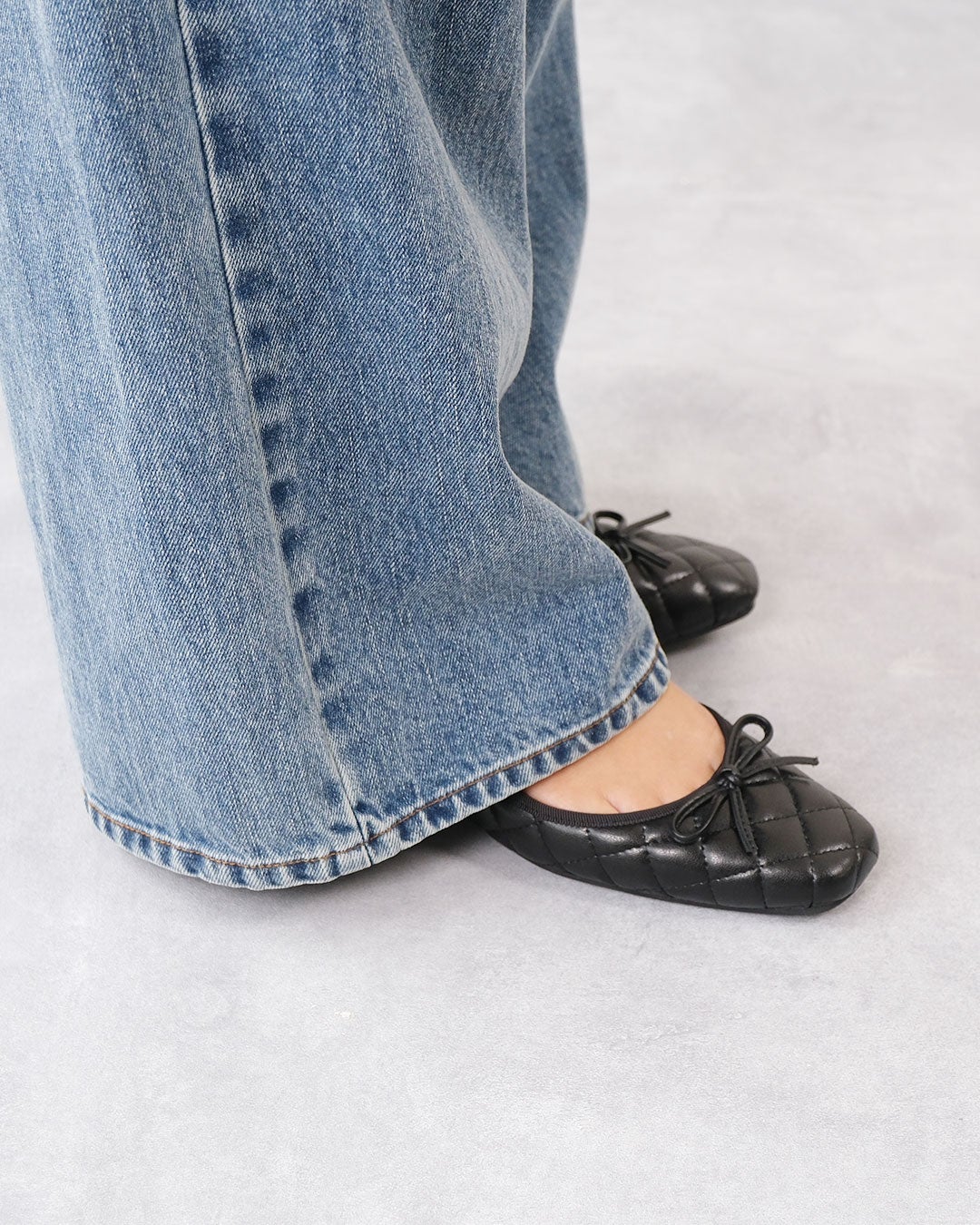 ワイドデニムパンツの裾から覗く、フロントにリボンをあしらったブラックのキルティングバレエシューズの着用足元拡大画像。
